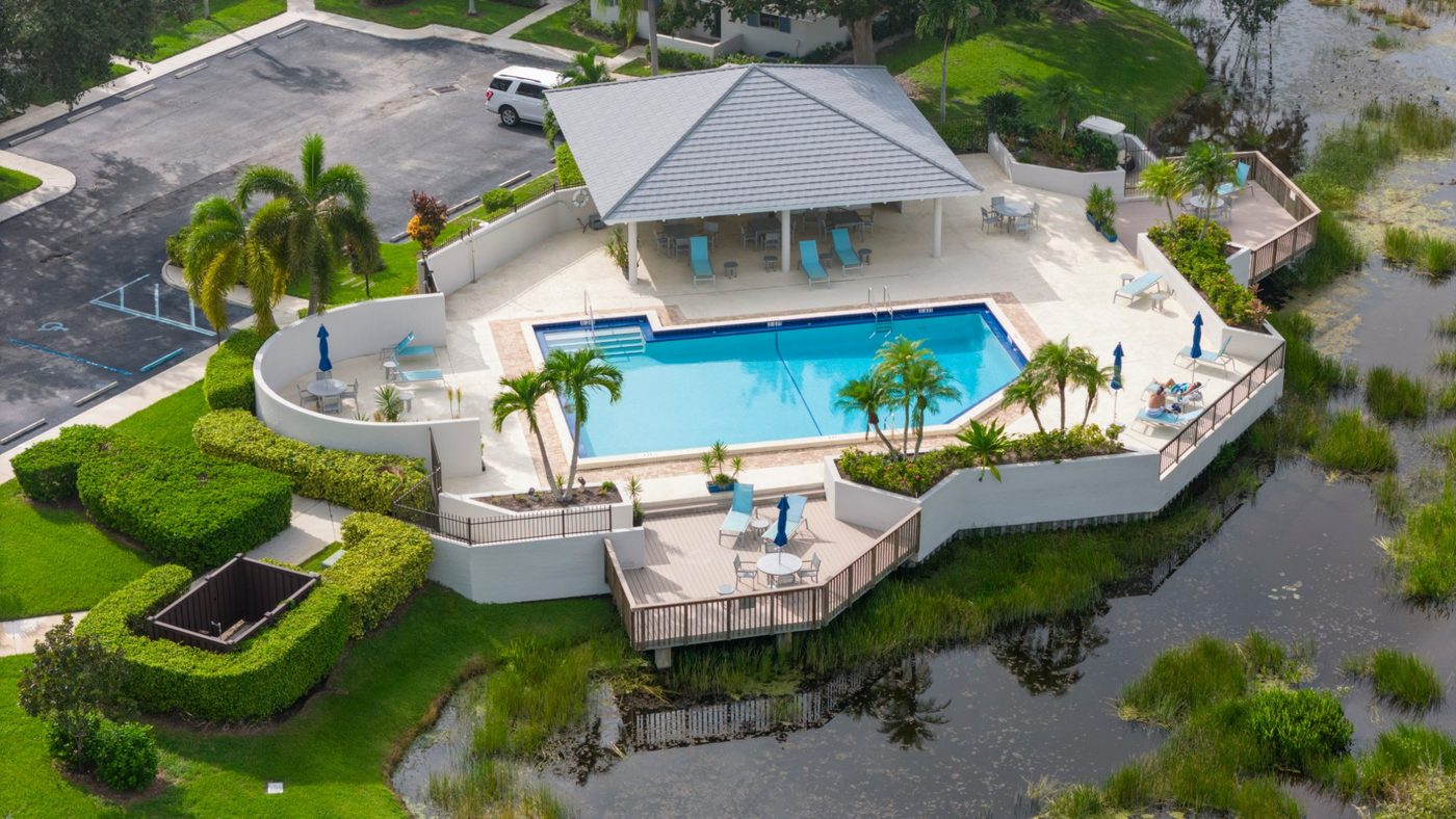 Aerial view of a community association amenity pool, a wide concrete deck, a covered pavilion and a raised perimeter wall